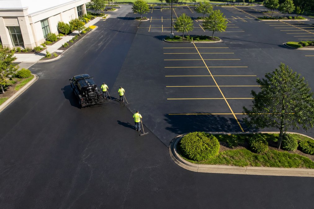 Aerial view of well-maintained commercial parking lot with clear markings, no visible cracks, and maintenance crew in safety vests performing sealcoating work under bright daylight