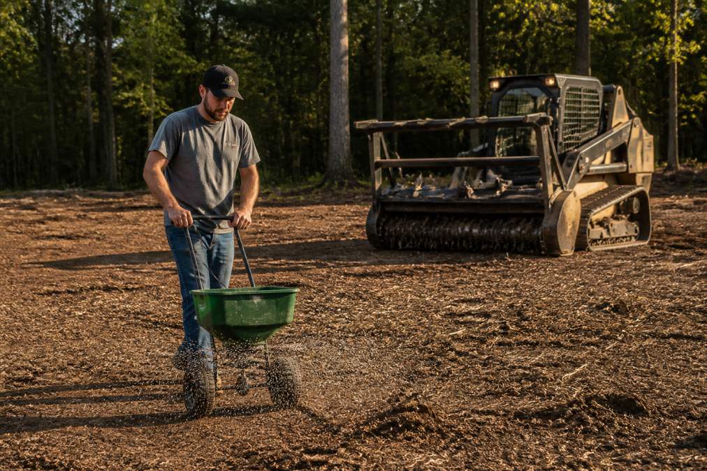 A person scattering grass seed over freshly cleared soil with a hand-cranked spreader, a large forestry mulcher parked on the bare dirt behind them, late afternoon sunlight casting long shadows