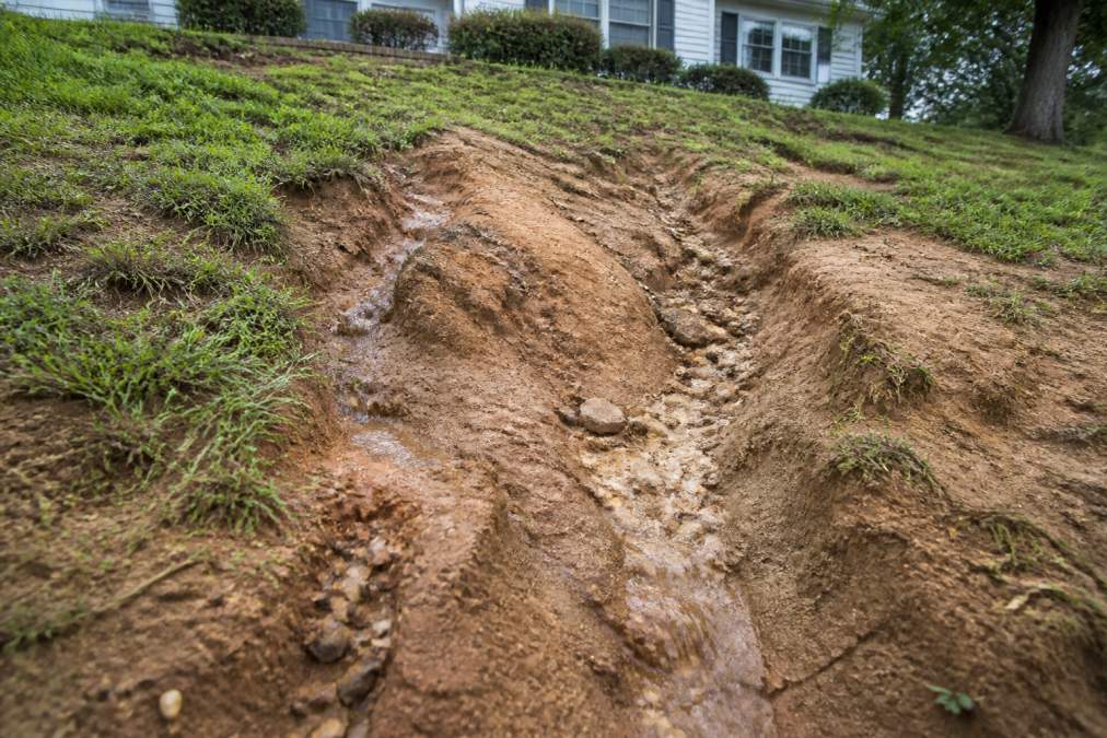 Close-up of eroded residential slope showing visible gullies, sediment displacement, and exposed subsoil with water runoff channels