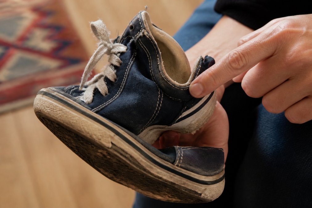 Close-up of a child's worn sneaker showing flattened tread, frayed laces, and loose heel counter, with a parent's hand pointing to areas of wear and concern