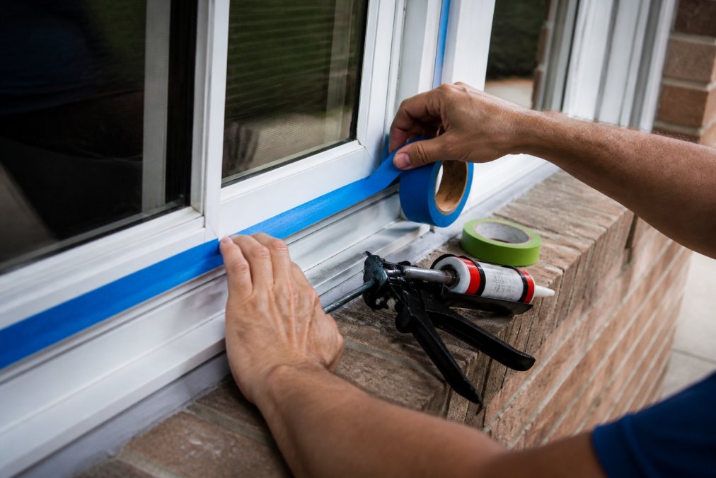 Hands applying waterproof tape to a window frame for preventing water damage during house pressure washing