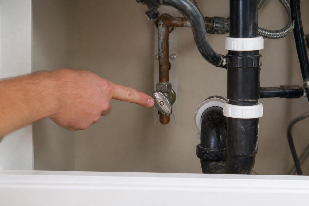 Close-up of a homeowner's hand pointing to a main water shutoff valve beneath a sink, showing the valve handle and surrounding pipes in clear detail