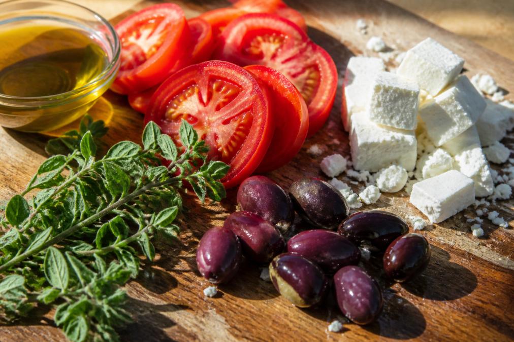 Close-up of fresh Greek ingredients arranged on a wooden cutting board: ripe red tomatoes with visible seeds, dark Kalamata olives, crumbled white feta cheese, fresh green oregano sprigs, and golden olive oil in a small glass bowl, with natural sunlight creating shadows across the surface