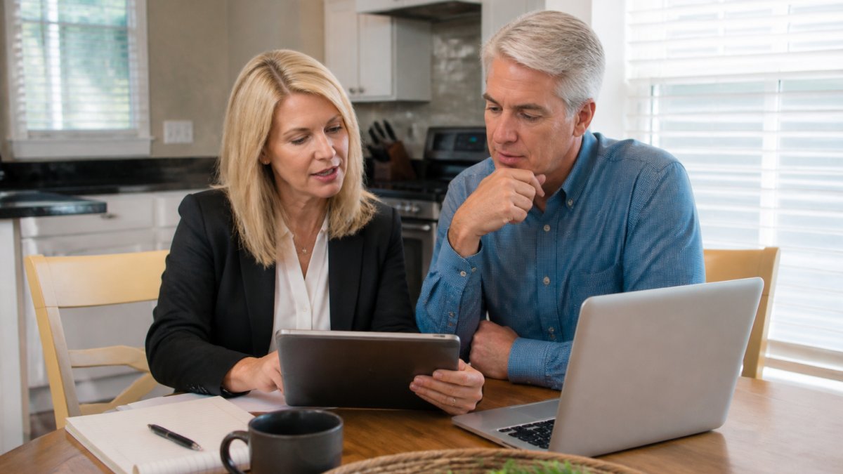 A real estate agent and homeowner reviewing comparable sales data on a tablet at a kitchen table, with a laptop and notepad nearby, soft morning light through the window