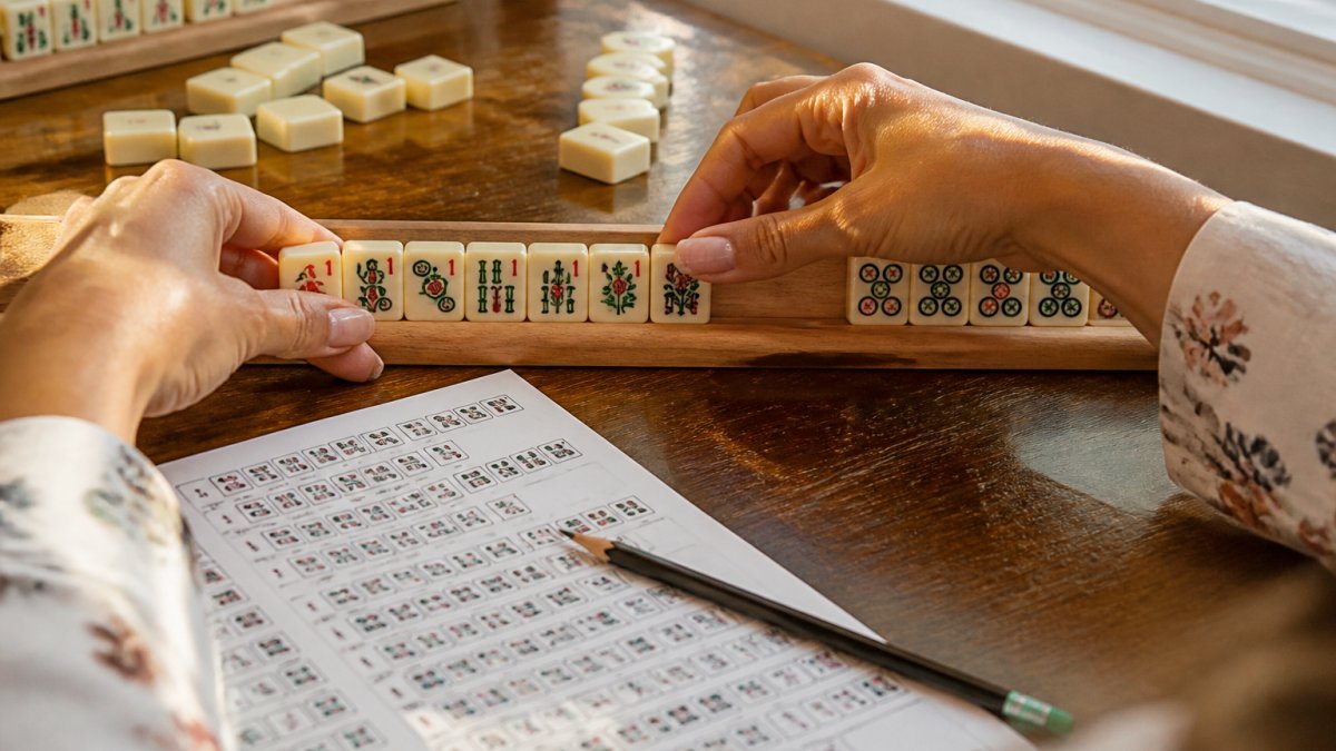 Close-up of a woman's hands arranging Mahjong tiles on a rack, with a printed hand guide and pencil nearby on a wooden table, warm afternoon light from a window