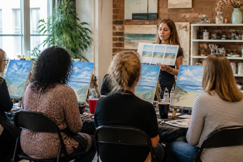 Art instructor holding printed painting instructions for a group of beginners in a bright paint and sip studio
