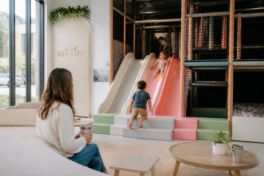 Bright, colorful indoor play space with children laughing and climbing on soft play structures while a parent watches from a nearby seating area with natural light streaming through large windows