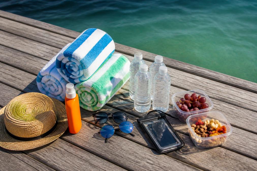 Overhead view of beach essentials laid out on a wooden dock: sunscreen, towels, snacks, water bottles, sunglasses, and a waterproof phone case, with calm Gulf waters in the background