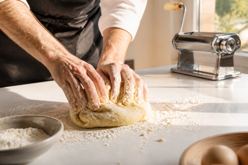 Close-up of hands kneading fresh pasta dough on a white kitchen bench, with semolina flour dusting the work surface and a stainless steel pasta machine visible in the background, natural afternoon light streaming across the counter