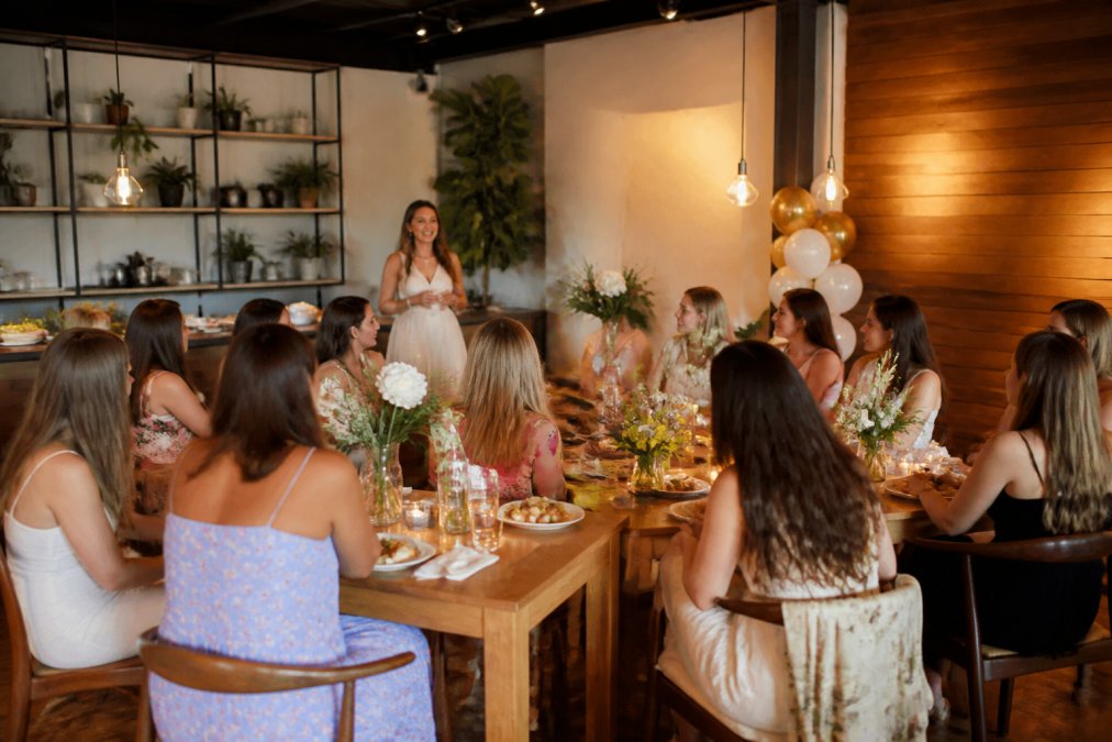 Small bridal shower celebration with guests seated around tables with fresh flowers and refreshments in a modern event space with warm ambient lighting