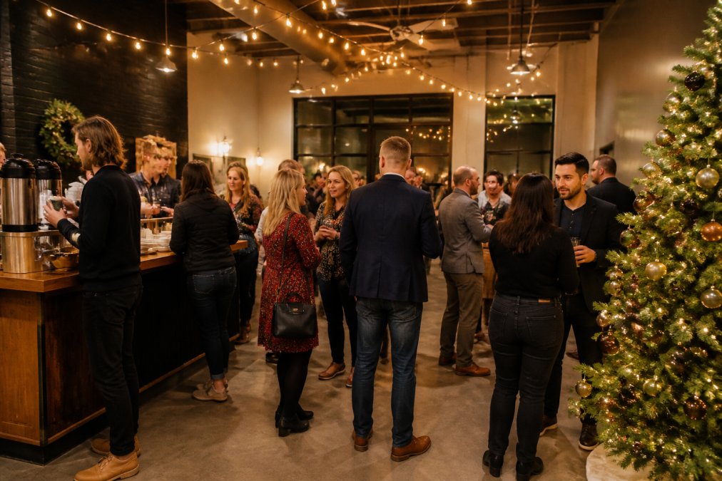 A festive holiday party in a warmly lit event space, string lights overhead, a decorated evergreen tree in the corner, and guests mingling near a coffee and catering bar