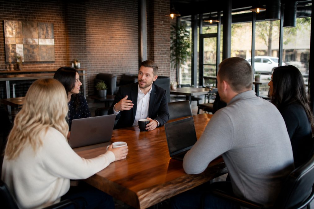 Small group of professionals gathered around a table in a modern, well-lit event space with coffee cups and laptops, engaged in conversation