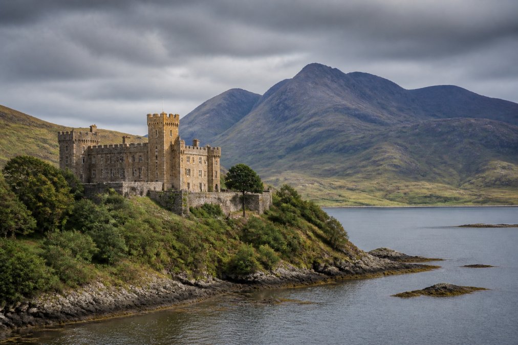 Panoramic view of Dunvegan Castle perched dramatically on the coastline with rugged mountains rising behind it under moody Highland sky, showcasing the iconic landscape that defines Skye's most famous landmarks