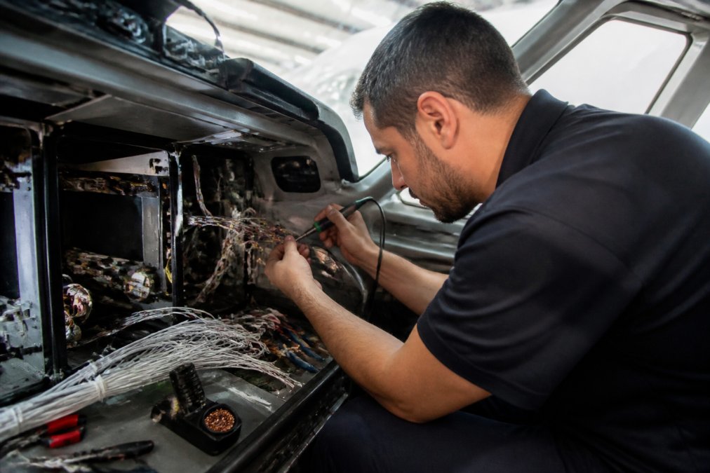 Professional aircraft technician carefully soldering electrical connections inside a complex avionics wiring harness while seated in the cockpit of a business jet, tools and wire bundles visible on the instrument panel, bright workshop lighting
