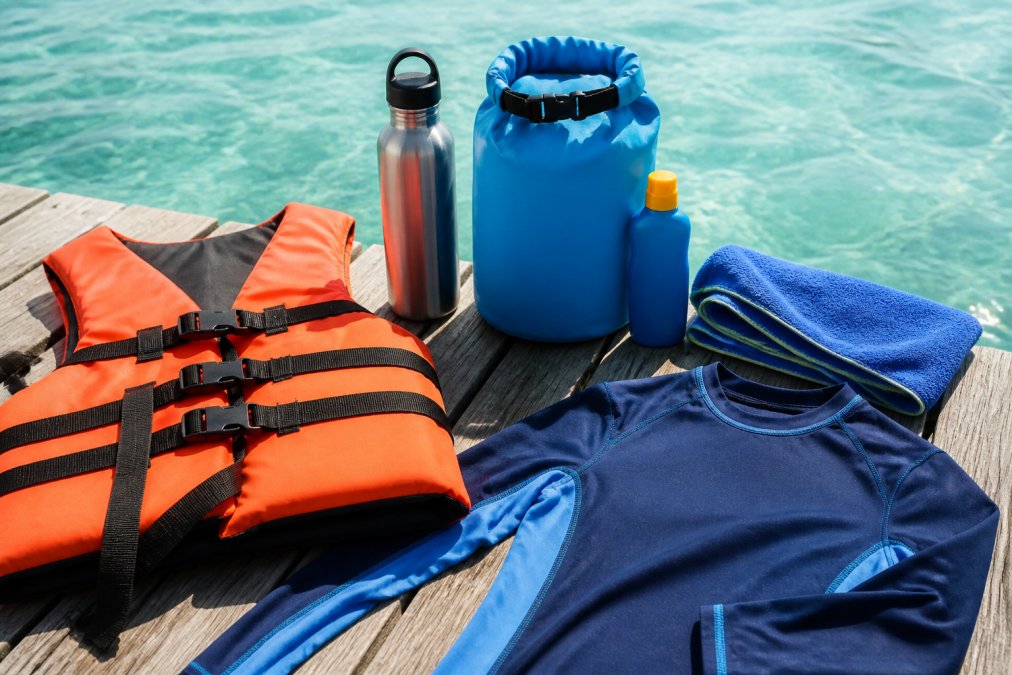 Flat-lay arrangement of jet ski essentials on a weathered wooden dock: sunscreen bottle, waterproof dry bag, microfiber towel, stainless steel water bottle, orange life jacket, and rash guard laid out with calm turquoise Gulf water visible in the background