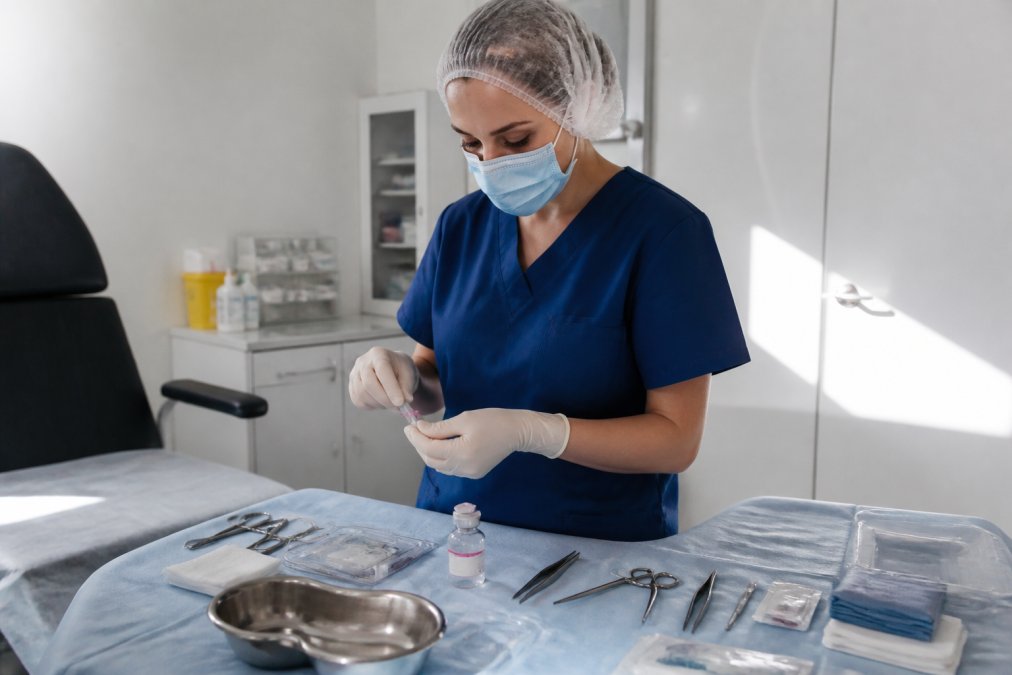Healthcare professional in modern private clinic preparing sterile instruments and local anaesthetic for minor surgical procedure, with bright overhead lighting and clean clinical environment