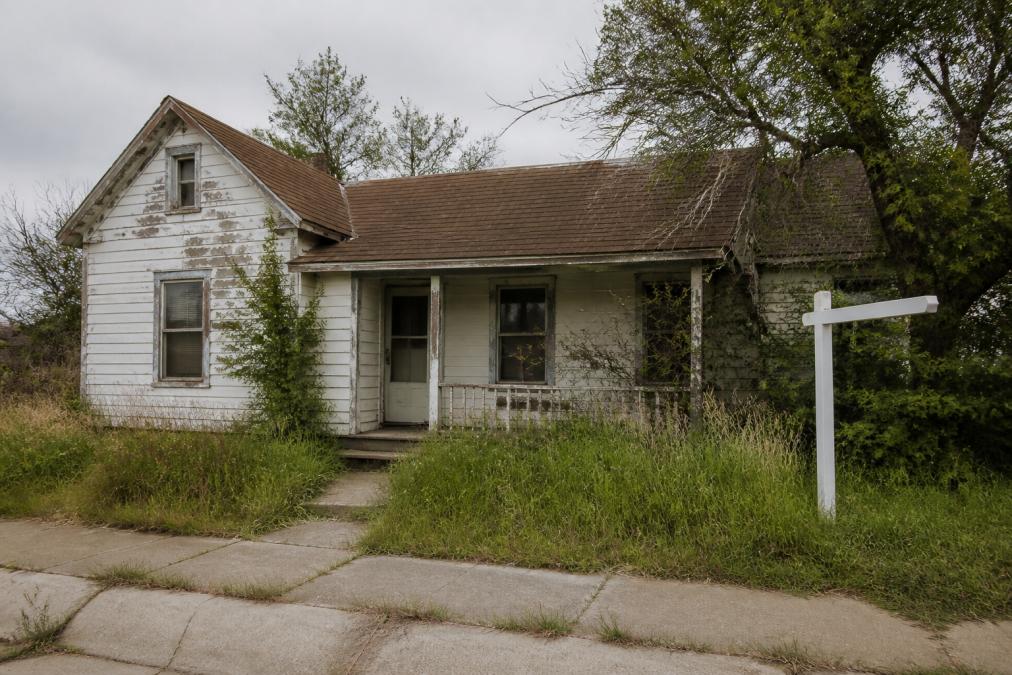 A distressed single-family home exterior with a 'For Sale' sign in the front yard, visible signs of deferred maintenance including peeling paint and overgrown landscaping, photographed on a cloudy day