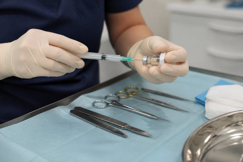 Medical professional's hands preparing sterile surgical instruments and local anaesthetic vial in a clinical setting with bright overhead lighting