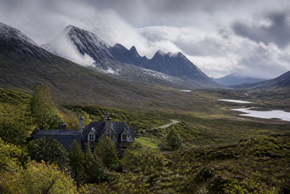 Dramatic Highland landscape showing rugged mountain peaks of the Cuillin range, wild moorland terrain, and a traditional stone lodge nestled in a misty valley below with lochs visible in the distance