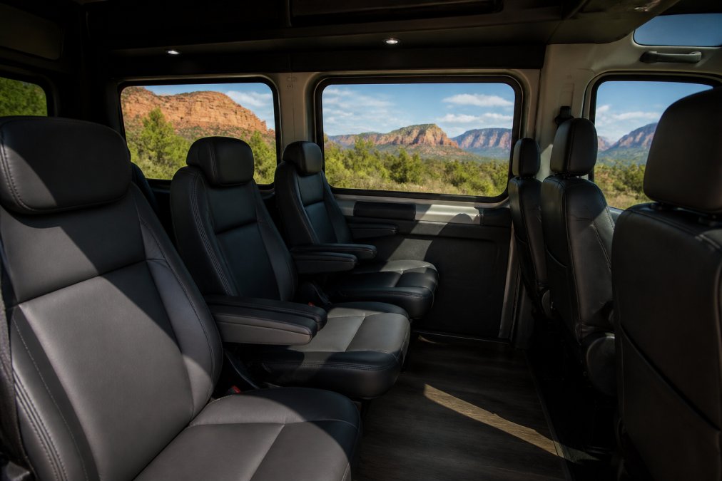 Interior of a comfortable Ford Transit van with premium seating and large windows, showing spacious legroom and Arizona landscape visible through the glass, with natural sunlight streaming in