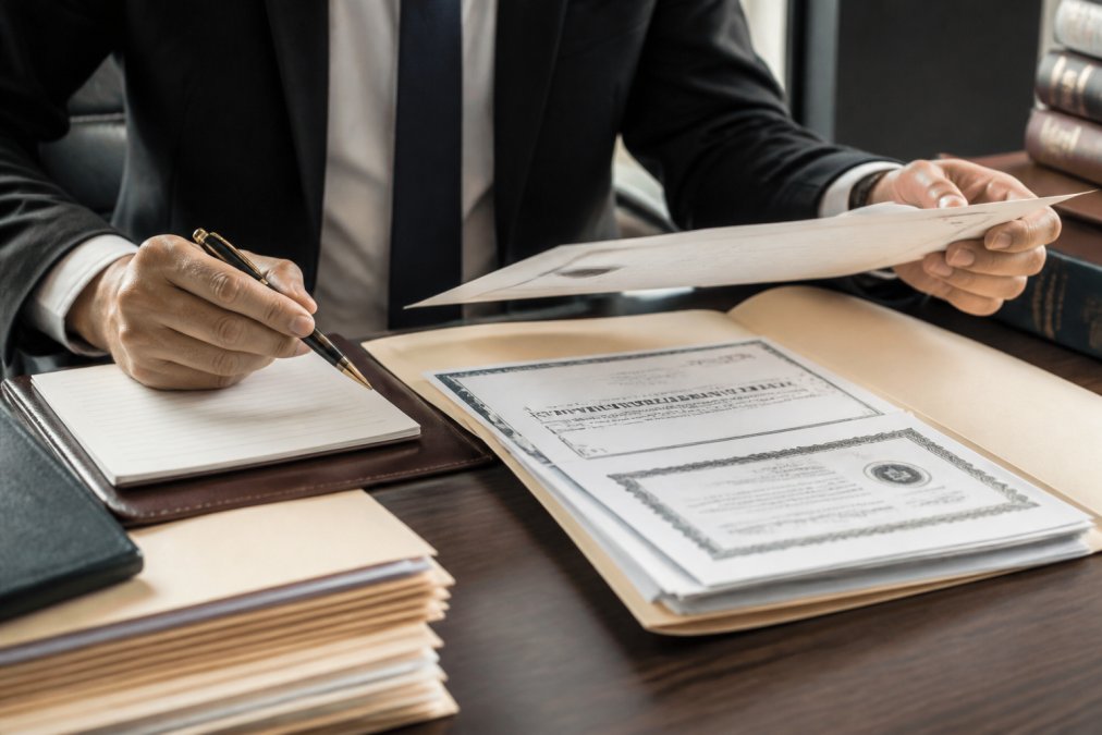 Close-up of a legal professional reviewing credentials and licensing documentation on a desk with a laptop and notepad, organized files and certification papers visible