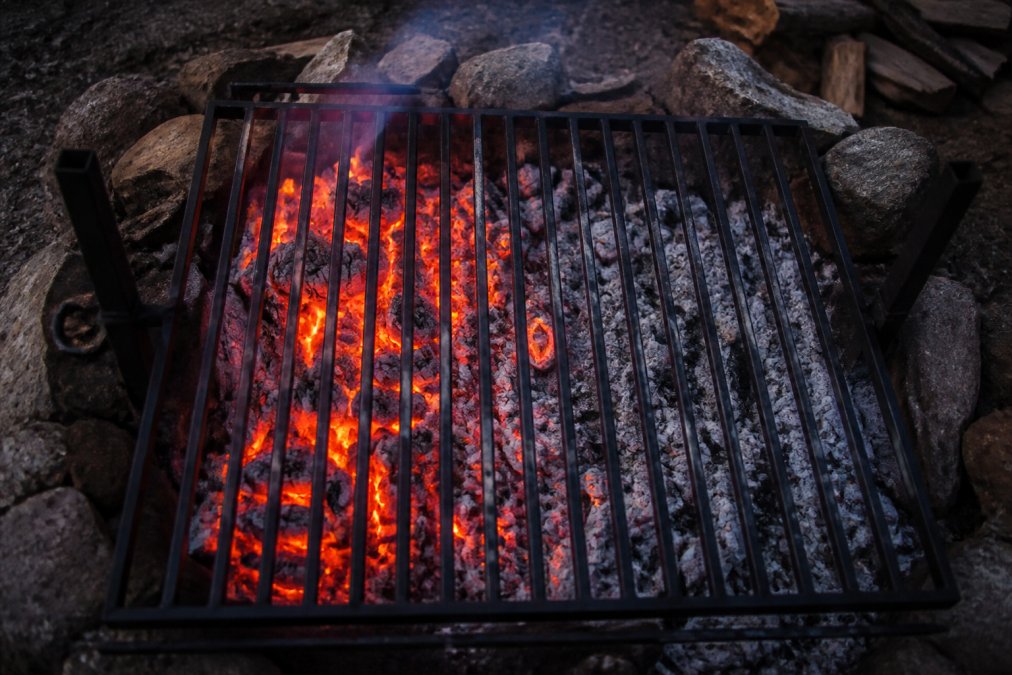 Overhead close-up of a campfire with a heavy cast iron grill grate positioned over glowing coals, showing a distinct gradient of bright orange embers on one side fading to ash-covered coals on the other, with smoke rising into dusk light
