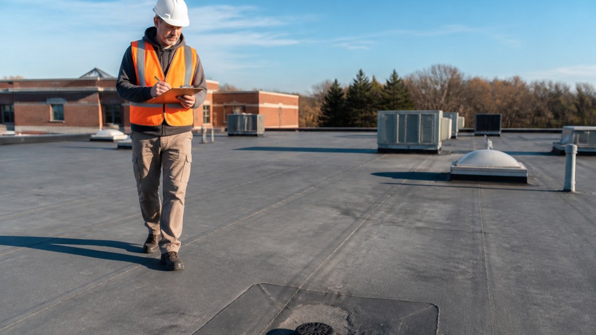 A facility director in a hard hat and safety vest walking a flat school roof with a clipboard, checking seams and drains on a clear morning