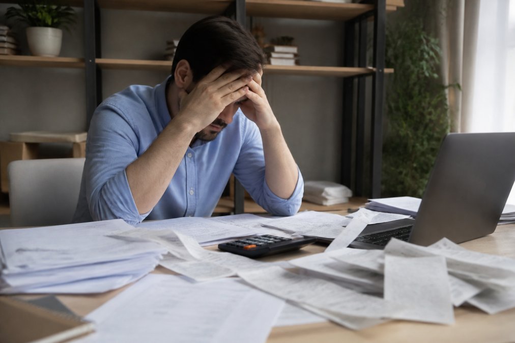 Stressed business owner sitting at a desk surrounded by tax documents and receipts, looking overwhelmed while staring at a computer screen with papers scattered across the desktop