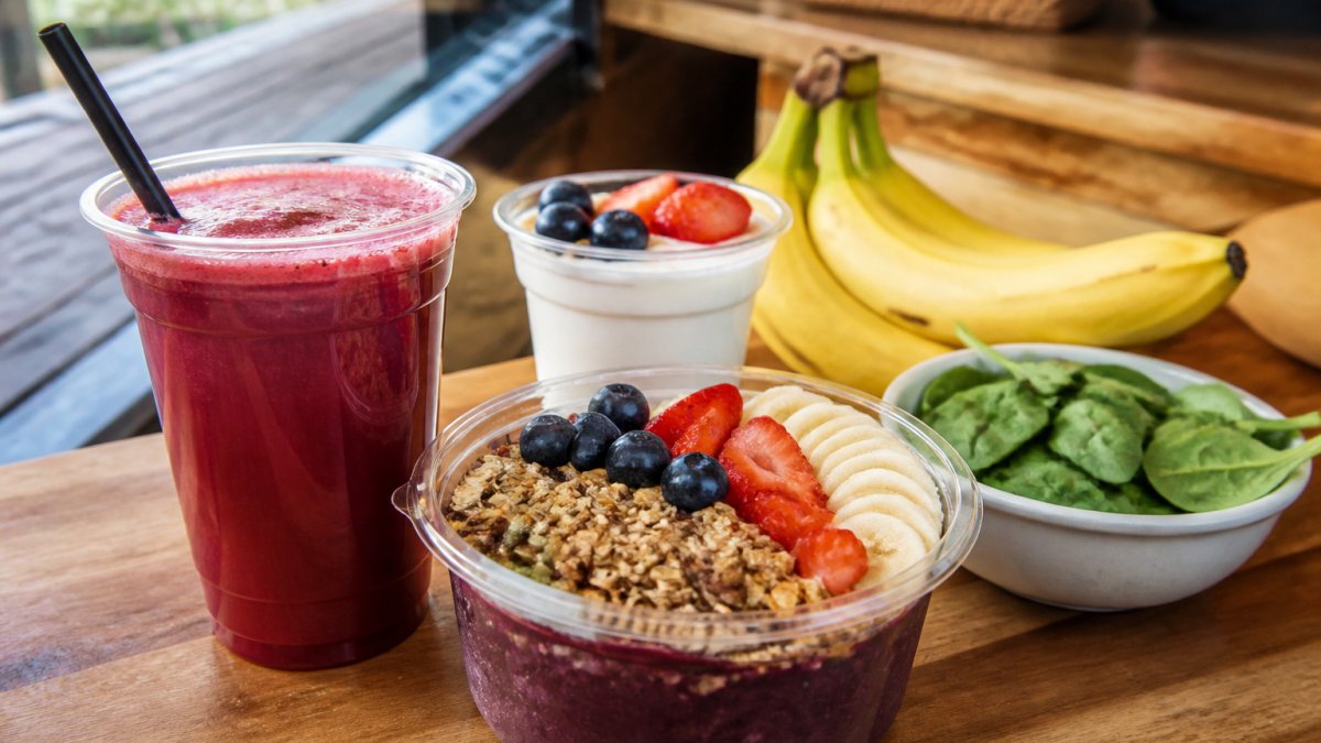 A colorful fresh-pressed juice and a smoothie bowl topped with granola on a wooden counter, with yogurt, bananas, and leafy greens visible nearby