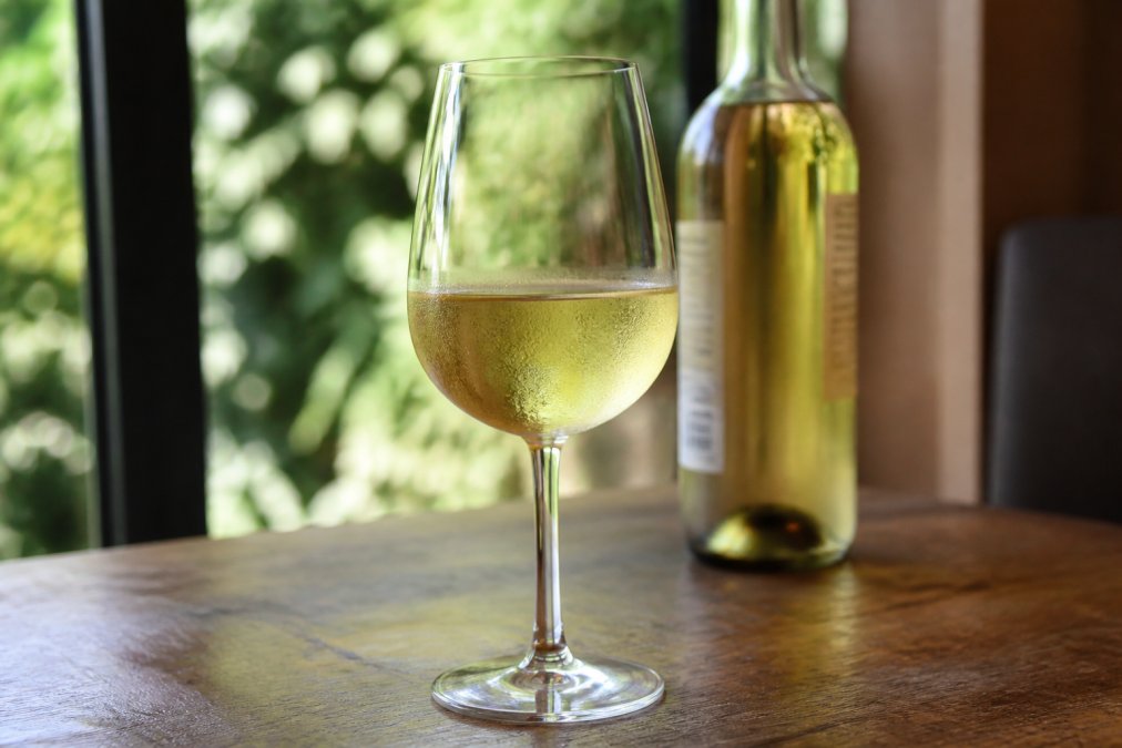 Close-up photograph of a chilled glass of pale alcohol-free white wine on a wooden table, with the bottle softly blurred behind it and natural daylight from a window