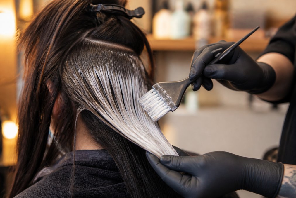 Close-up of a colorist's hands holding a tint brush mid-application, feathered bristles loaded with lightener sweeping through a sectioned panel of hair, warm salon lighting
