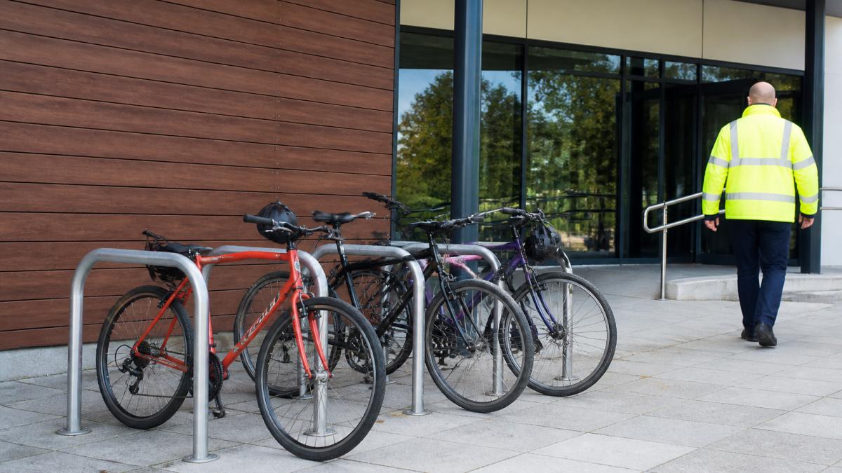 A galvanised Sheffield-style cycle rack installed outside a council leisure centre, with several bicycles parked and a person in a high-vis jacket walking past