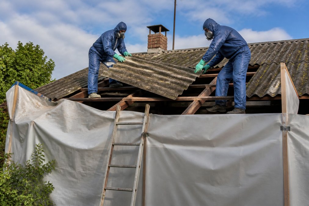 Dva pracovníci v ochranných oblecích, respirátorech a rukavicích demontují eternitové šablony ze střechy rodinného domu, kolem je zajištěno staveniště se zakrytými plochami a plastovými fóiemi, denní světlo, dokumentární styl