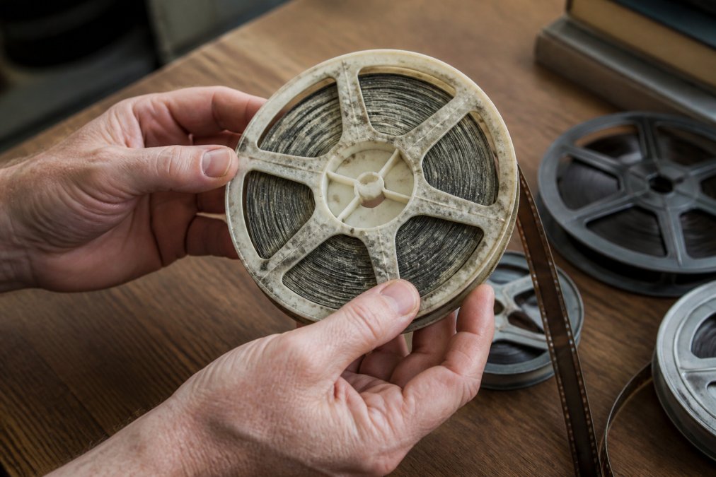 Professional hands carefully holding and examining old 8mm film reels, showing signs of dust and aging on the film surface, with natural light highlighting degradation patterns