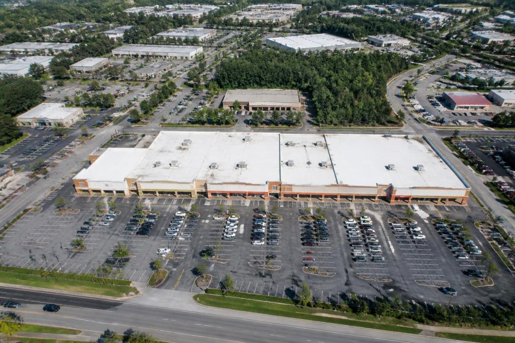 Aerial view of a commercial property complex showing roofline, parking lot, building footprint, and surrounding businesses and infrastructure from above in clear daylight