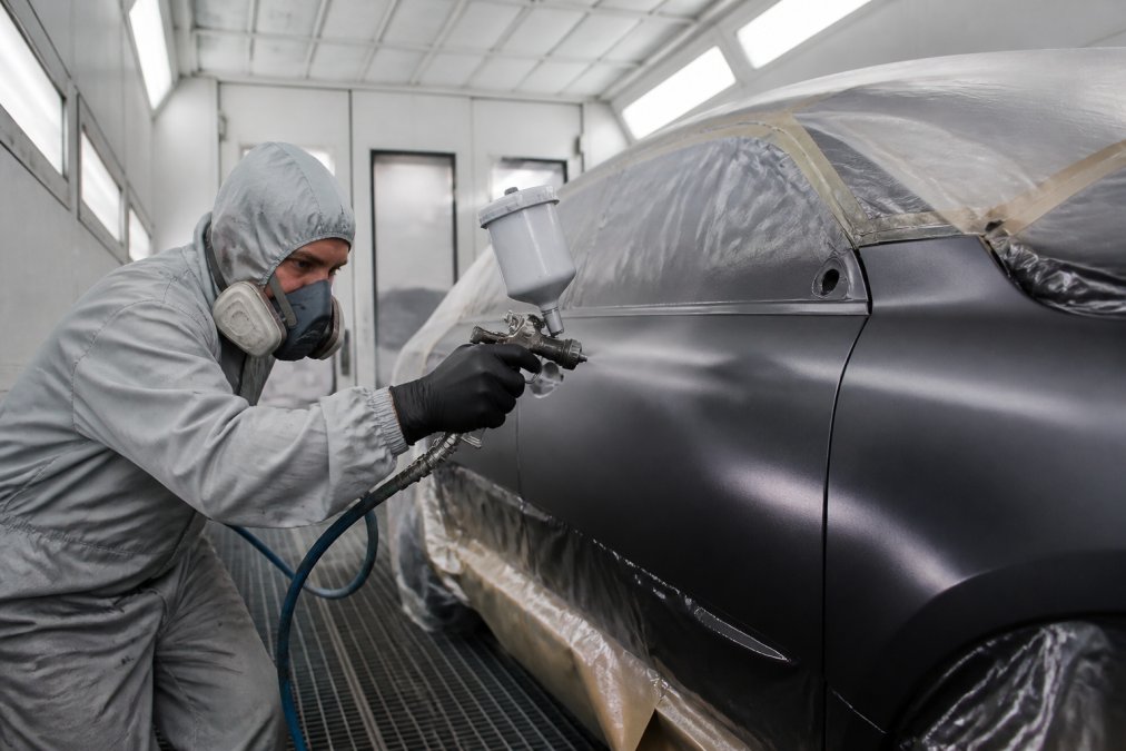 Certified technician in professional paint booth spraying basecoat and clear coat on vehicle panel with spray gun, proper ventilation system and protective equipment visible