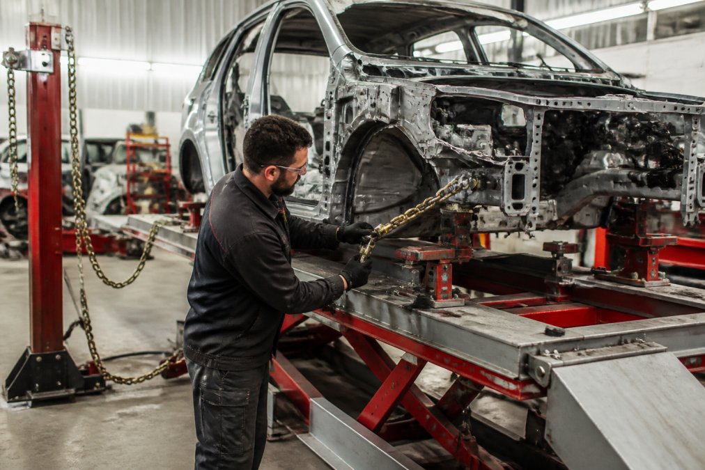 Professional technician working on a vehicle positioned on a drive-on frame machine in a modern collision repair shop, with hydraulic lifting systems visible and bright overhead LED lighting illuminating the work area