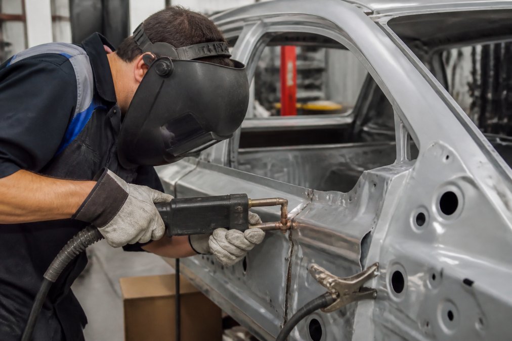 Professional technician in safety gear performing precision spot welding on an aluminum vehicle panel using specialized welding equipment, with close-up focus on the weld joint and the tool's positioning