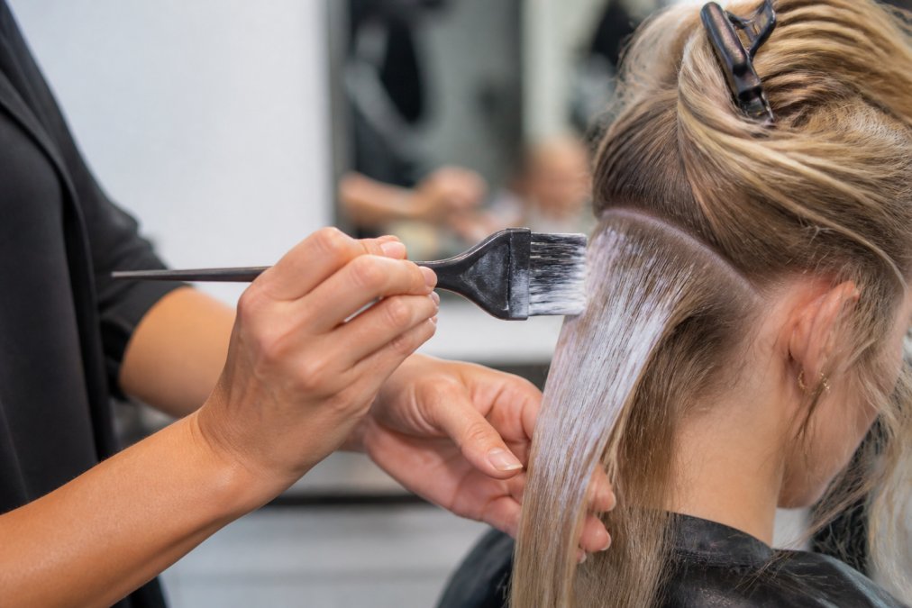 Close-up of a colorist's hands holding a feathered balayage brush applying lightener to a sectioned strand of hair in a bright salon