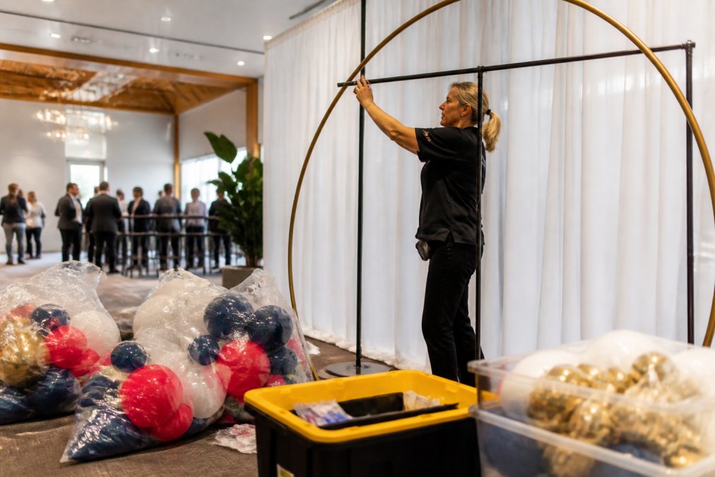 Professional balloon artist securing freestanding demi-arch structure to backdrop stand at corporate event venue with attendees in background, focused lighting on installation work