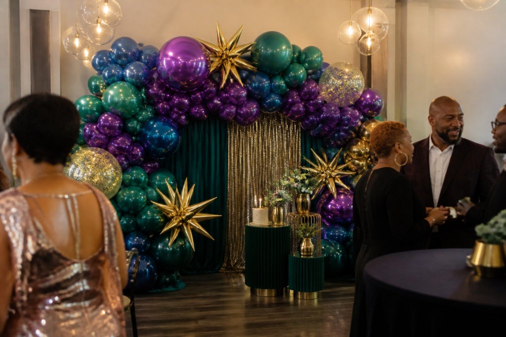 Professional balloon backdrop featuring organic garland design with coordinated latex and foil balloons in jewel tones, displayed at a birthday venue with guests mingling in foreground, warm ambient lighting