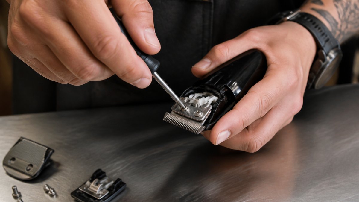Professional barber's hands adjusting clipper blade tension with precision screwdriver tool, showing proper hand positioning and alignment technique on stainless steel work surface