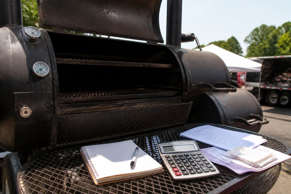 Close-up of a large heavy-gauge steel offset smoker with visible cooking chamber, firebox, and thick welded seams, positioned outdoors at a competition BBQ event in afternoon sunlight