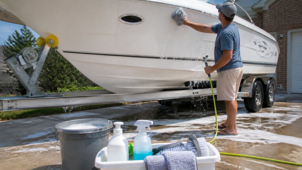 Boat owner washing the white fiberglass hull of a boat in a sunny driveway using a soapy mitt and water hose