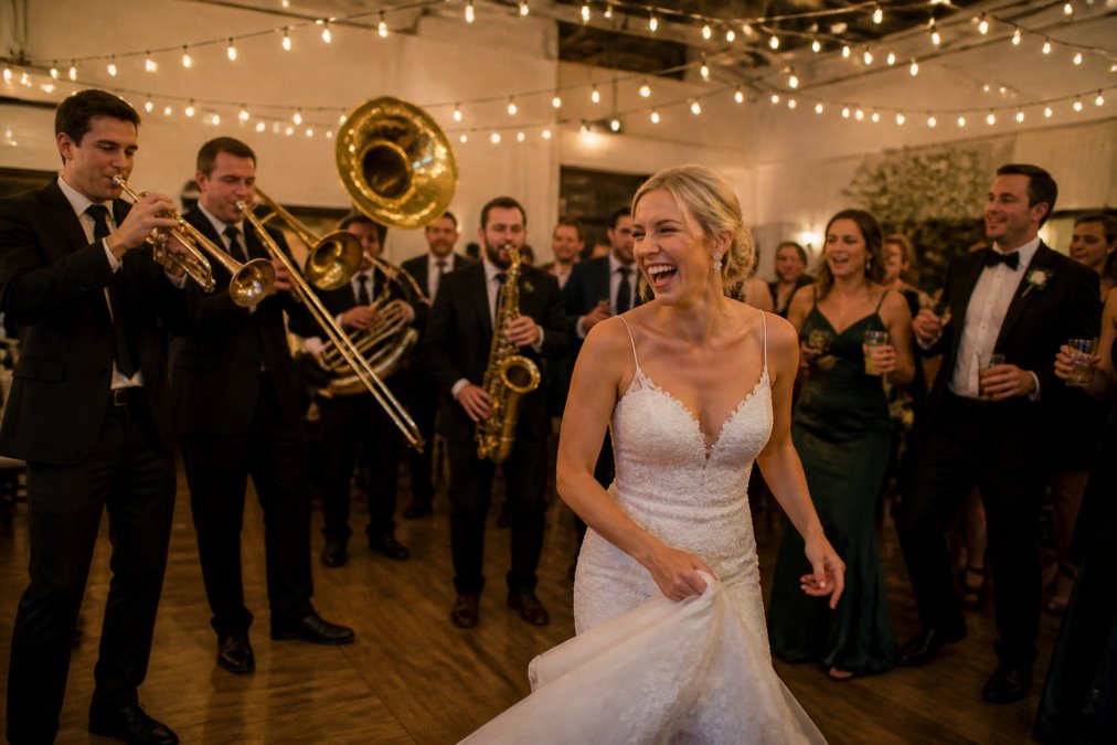 A five-piece brass band in matching suits performing at an elegant wedding reception, guests dancing on a hardwood floor beneath warm string lights, a bride in a white gown laughing in the foreground