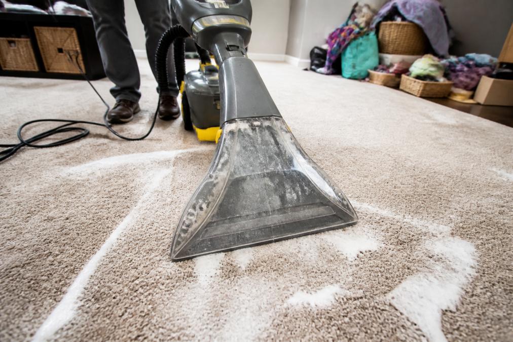 Close-up of a rental carpet cleaning machine being pushed over a damp residential carpet, with visible soapy suds and wet residue left behind in the carpet pile under bright indoor lighting
