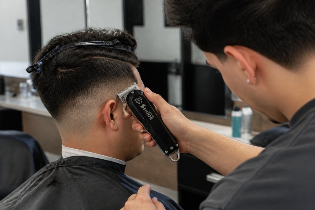 Apprentice barber demonstrating proper clipper grip and hand position during a fade cut, showing correct wrist angle and shoulder alignment with natural studio lighting