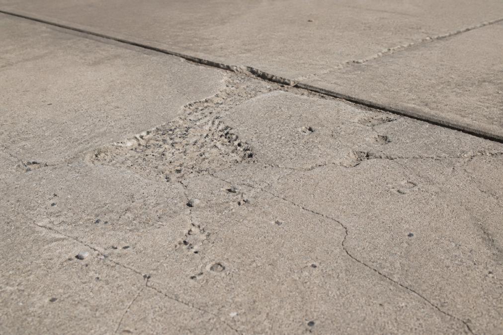 Close-up of a damaged concrete driveway surface showing visible spalling, hairline cracks, and surface pitting in natural daylight, with weathered concrete texture clearly visible