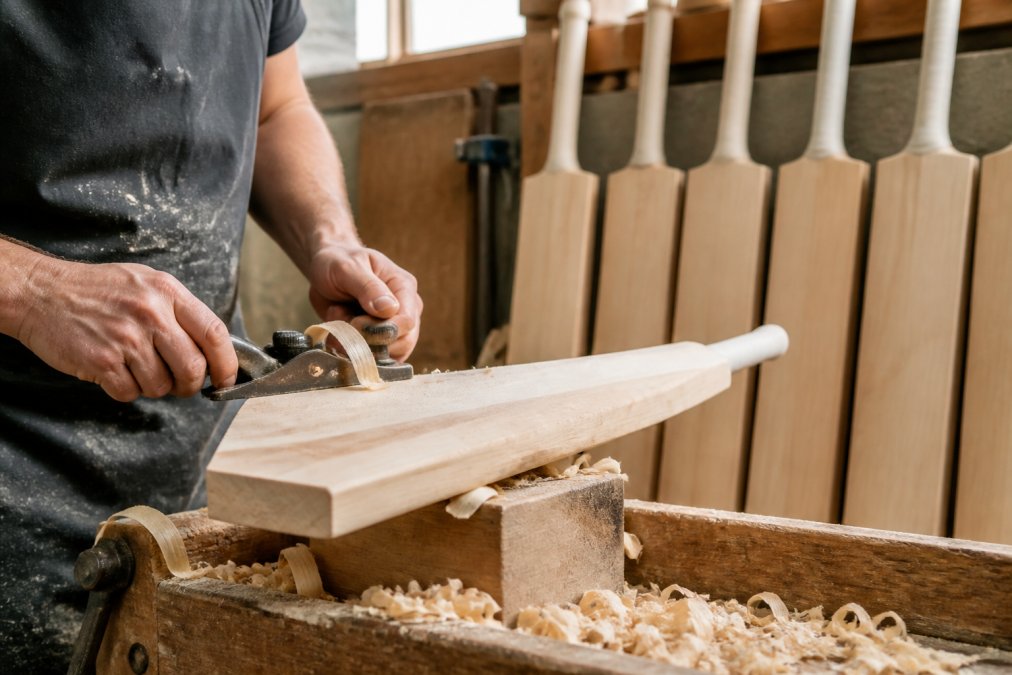 A craftsman's hands shaping a cricket bat with a spokeshave in a bright workshop, wood shavings on the bench and finished bats lined up behind