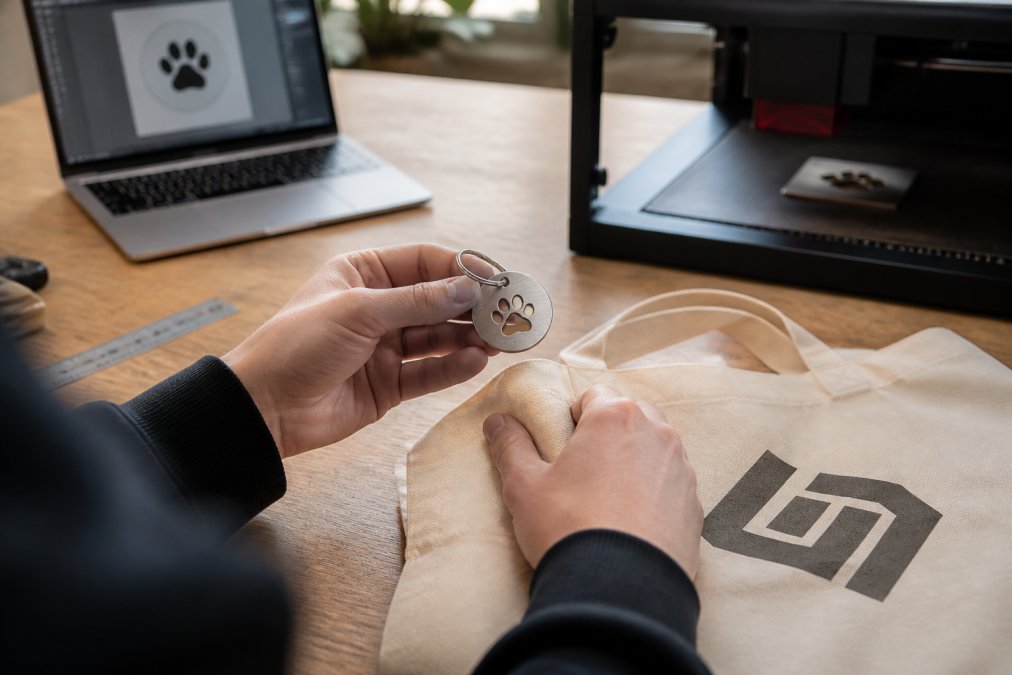 Close-up of a person's hands holding a newly laser-engraved metal pet ID tag and a branded tote bag on a clean workbench, with a laptop showing a design file in the background
